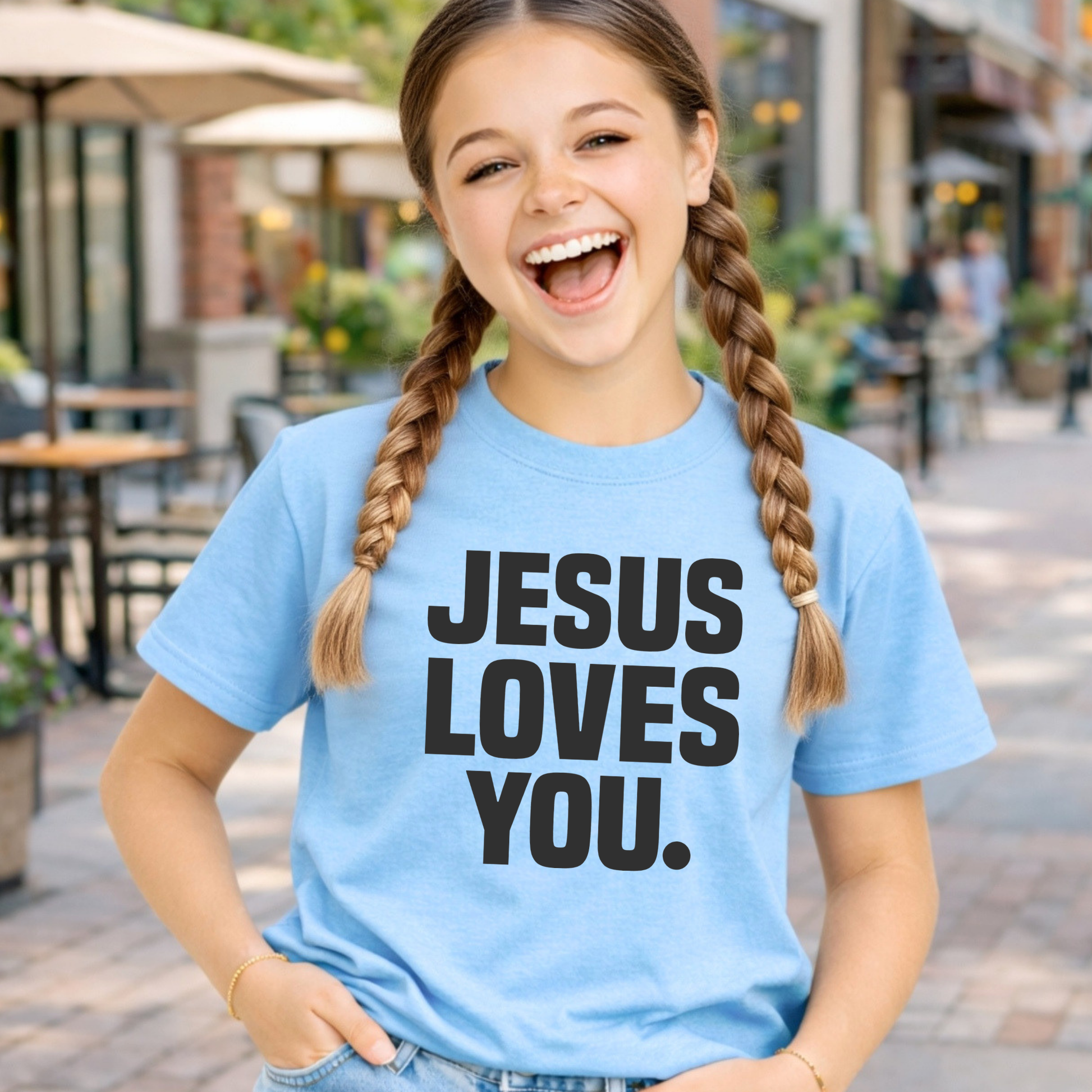 Young girl wearing a blue t-shirt with 'Jesus Loves You' text, standing outdoors.