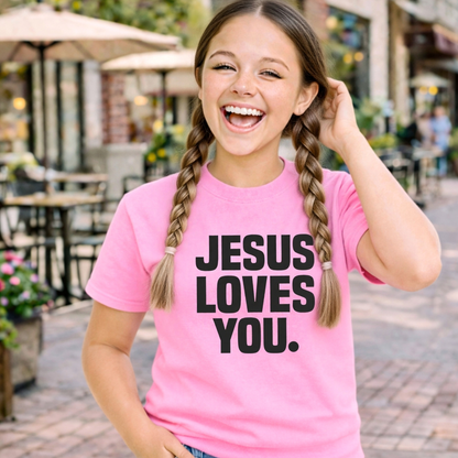 Young girl wearing a pink t-shirt with 'Jesus Loves You' text, standing indoors.
