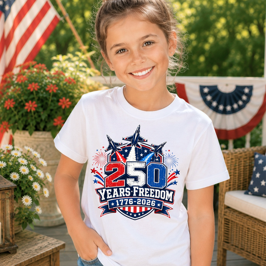 Young girl wearing a white t-shirt with a 250 Years Freedom design, standing outdoors with American flags and flowers in the background.