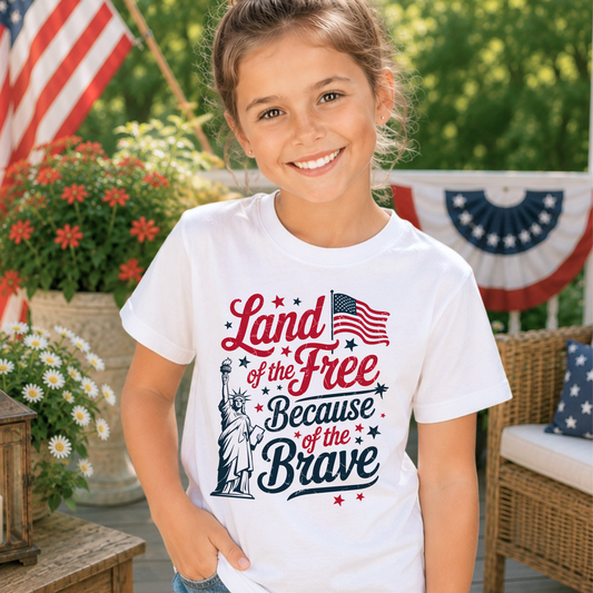 Young girl wearing a white t-shirt with a patriotic design, standing outdoors with an American flag and flowers in the background.