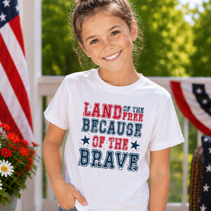 Young girl wearing a white t-shirt with patriotic text, standing outdoors with American flags and flowers in the background.