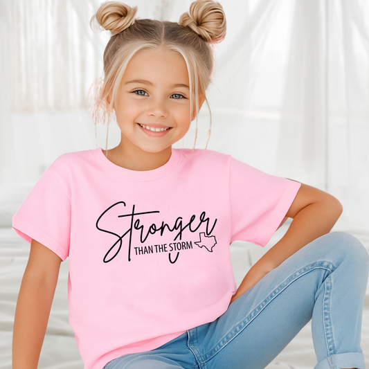 Young girl wearing light pink “Stronger Than the Storm” Texas kids T-shirt, smiling in bright room.

