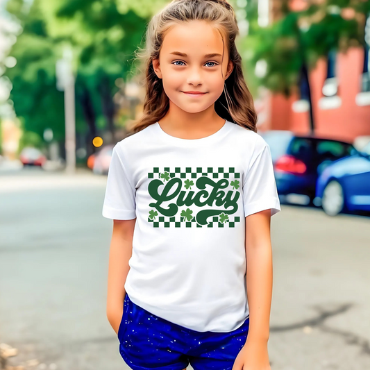 White T-shirt with 'Lucky' in green cursive, surrounded by shamrocks and a checkered pattern – perfect for St. Patrick’s Day.