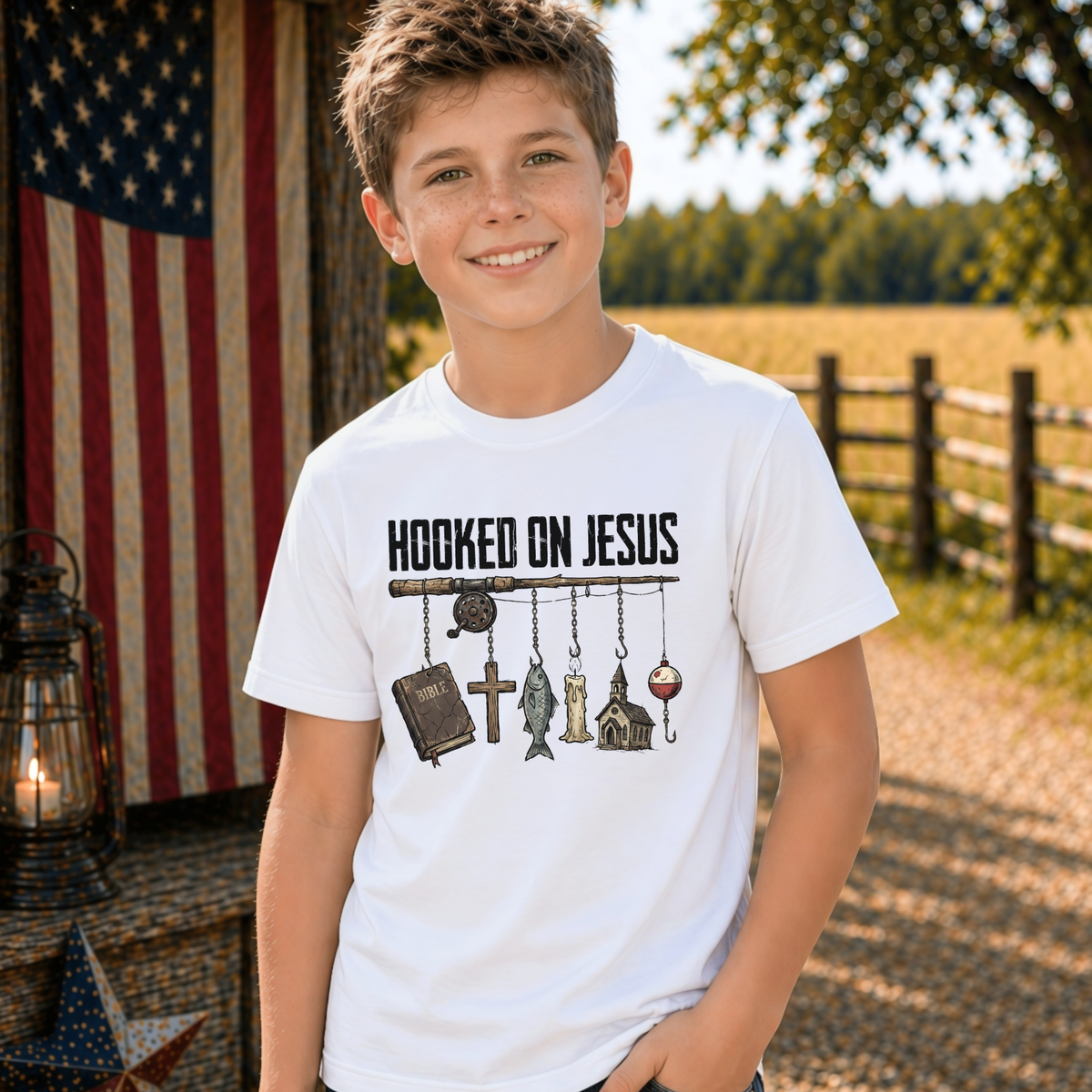 Boy wearing a white youth Hooked On Jesus T-shirt in a rustic outdoor setting with an American flag, lantern, and barn star.