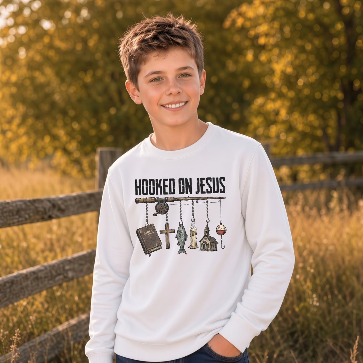 Boy wearing a white youth Hooked On Jesus sweatshirt beside a wooden fence in a sunny field.