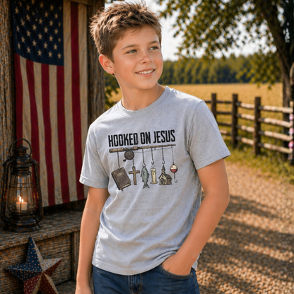 Boy wearing a sports gray youth Hooked On Jesus T-shirt in a rustic outdoor setting with an American flag, lantern, and barn star.