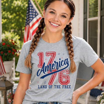 Girl wearing a gray t-shirt with '1776 America Land of the Free' design on a porch.