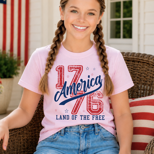 Young girl wearing a pink t-shirt with '1776 America Land of the Free' design, sitting on a porch with an American flag in the background.