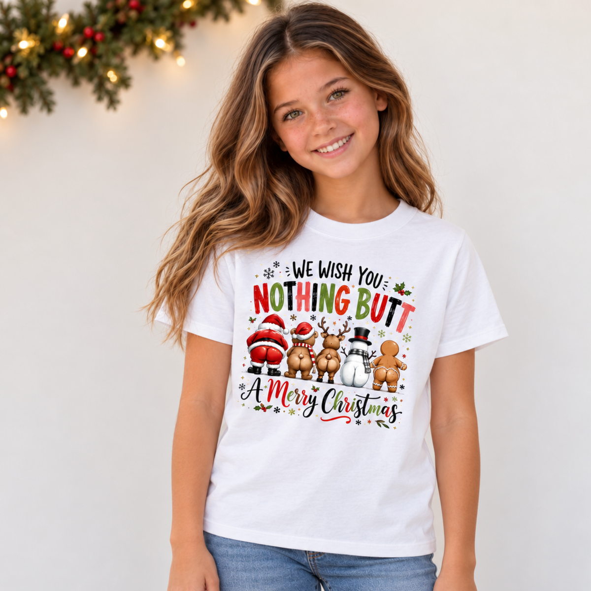 Youth model wearing a white T-shirt with We Wish You Nothing Butt a Merry Christmas design with festive characters shown from behind beneath simple Christmas greenery