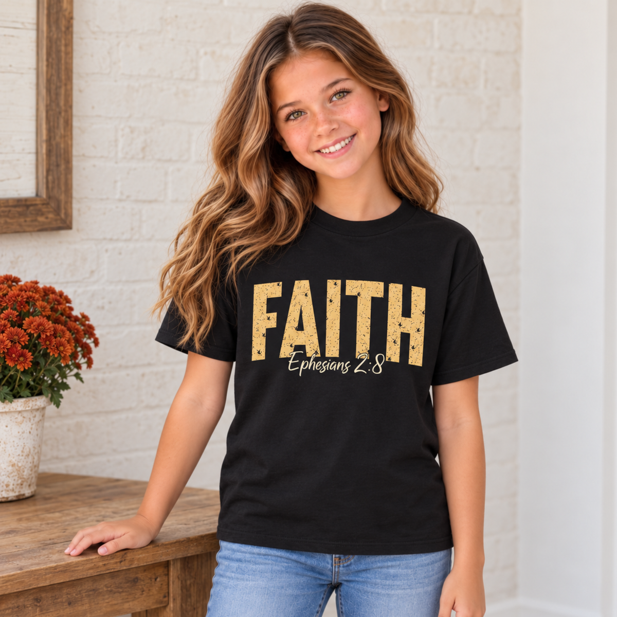 Young model wearing a black youth T-shirt with distressed gold FAITH lettering and white Ephesians 2:8 script beside a rustic table and orange fall mums.
