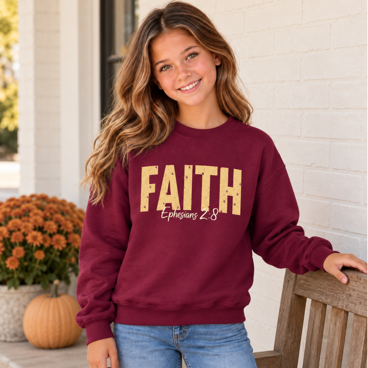 Young model wearing a maroon youth crewneck sweatshirt with distressed gold FAITH text and white Ephesians 2:8 script beside a porch bench, pumpkin, and fall mums.