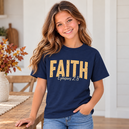 Young model wearing a navy youth T-shirt featuring distressed gold FAITH lettering and white Ephesians 2:8 script beside a farmhouse table with autumn leaves.