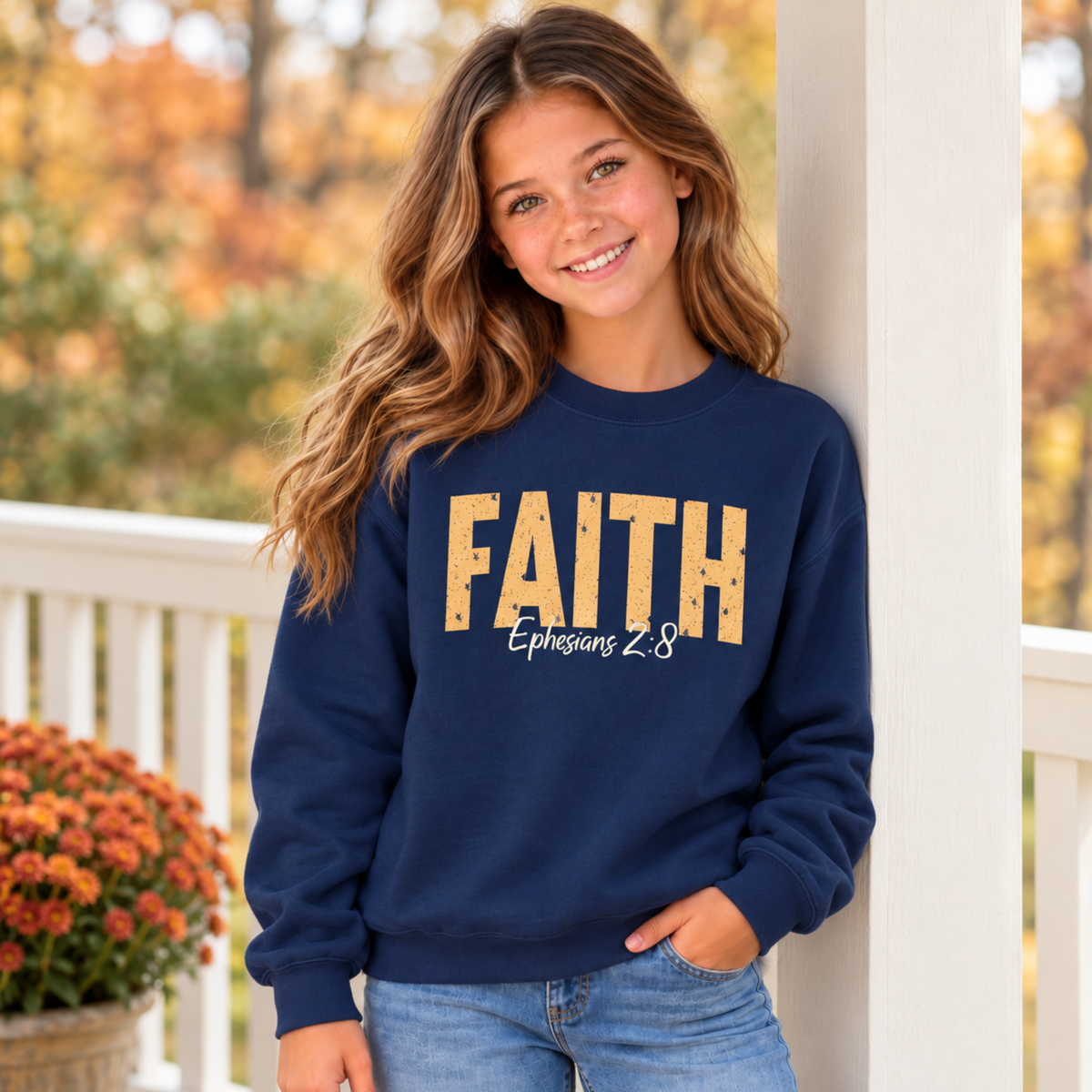 Young model wearing a navy youth crewneck sweatshirt with distressed gold FAITH text and white Ephesians 2:8 script on a porch with colorful fall trees and mums.
