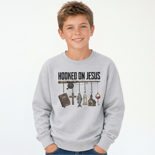 Smiling boy wearing a sports gray youth sweatshirt with a Hooked On Jesus fishing rod graphic featuring a Bible, cross, fish, church, and bobber.