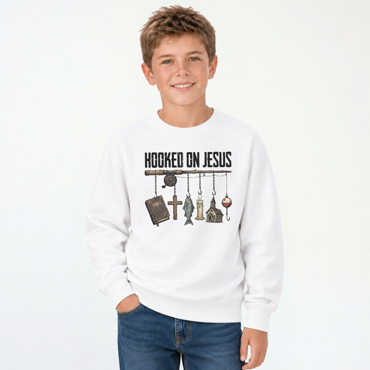 Smiling boy wearing a white youth sweatshirt with a Hooked On Jesus fishing rod graphic featuring a Bible, cross, fish, church, and bobber.