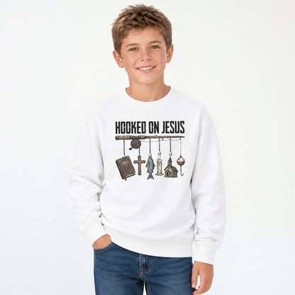 Smiling boy wearing a white youth sweatshirt with a Hooked On Jesus fishing rod graphic featuring a Bible, cross, fish, church, and bobber.