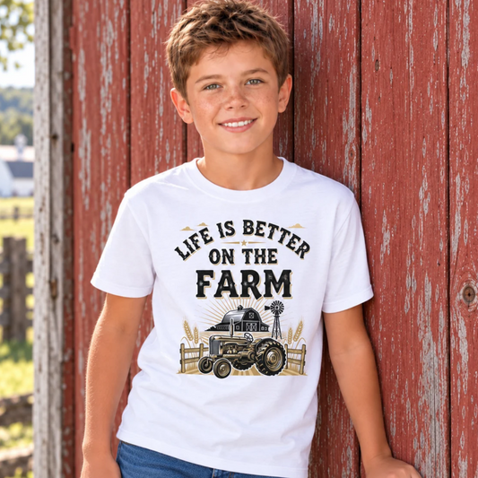 Boy wearing a white youth Life Is Better on the Farm T-shirt with tractor, barn, and windmill graphic against a weathered red barn wall.