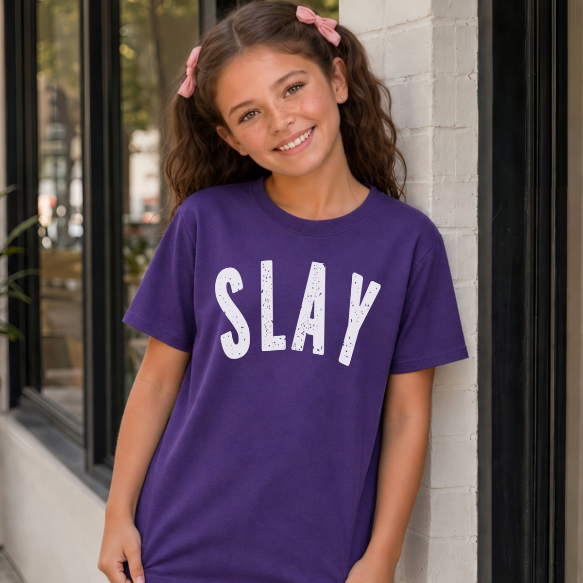 Young girl wearing a purple short-sleeve SLAY T-shirt with bold white distressed lettering outside a modern storefront.