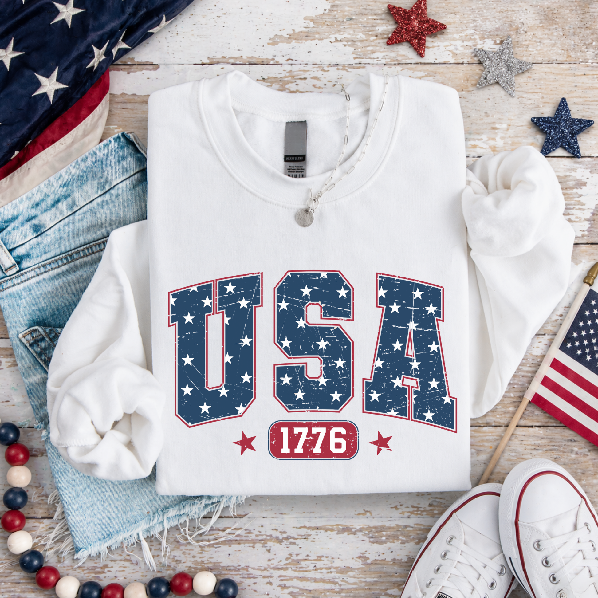 White sweatshirt with 'USA 1776' design on a wooden surface with American flags and stars.