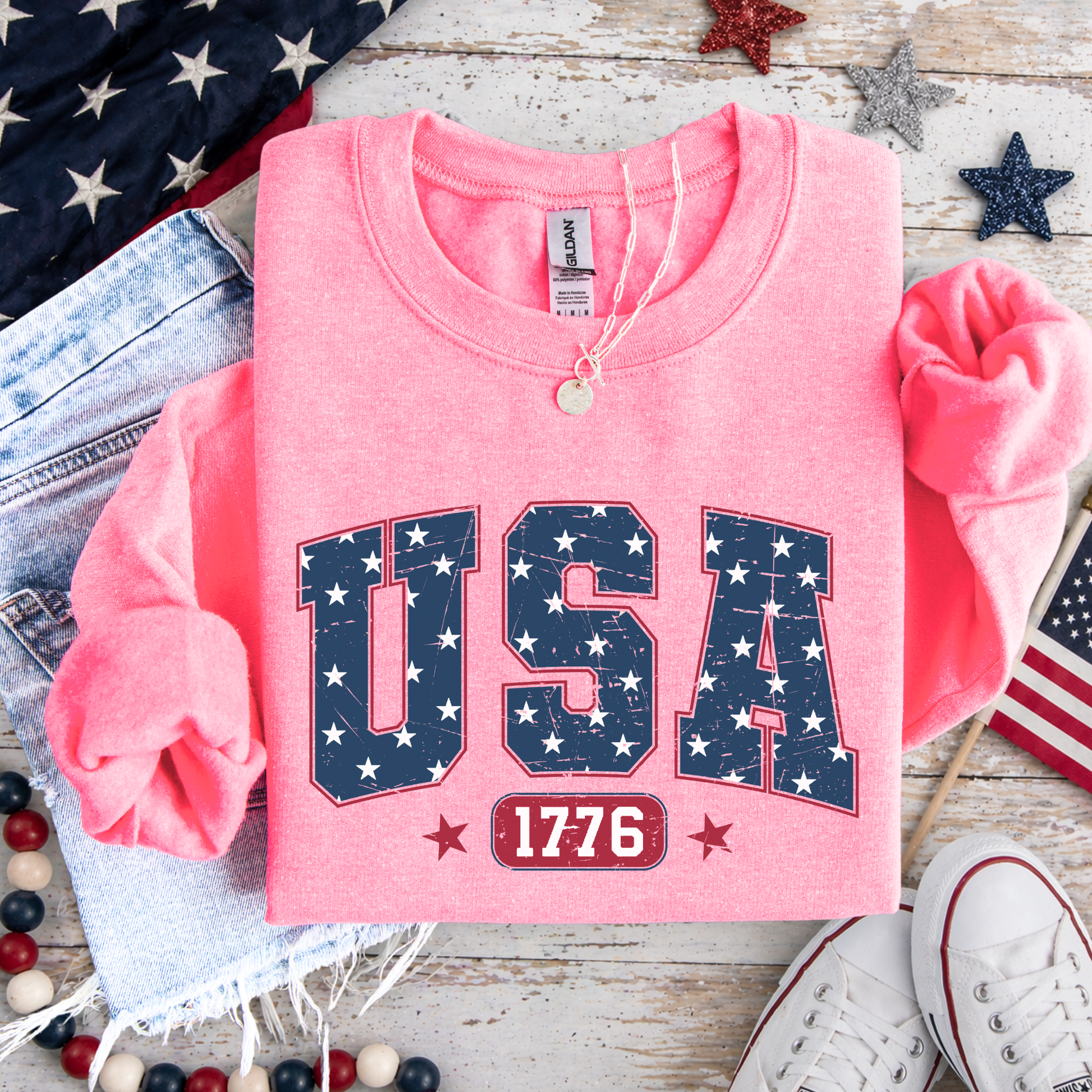 Pink sweatshirt with 'USA 1776' design on a wooden surface with American flags and shoes.