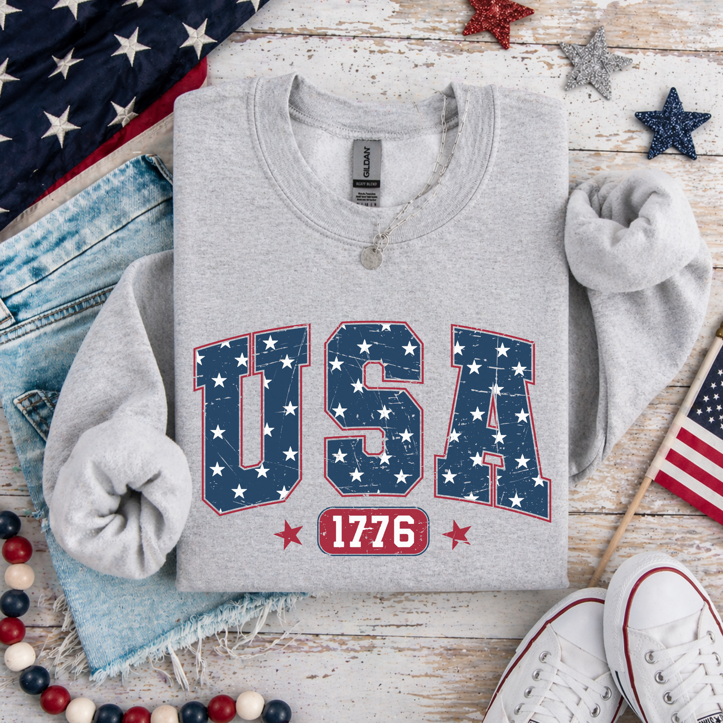 Gray sweatshirt with 'USA 1776' design on a wooden surface with American flags and jeans.