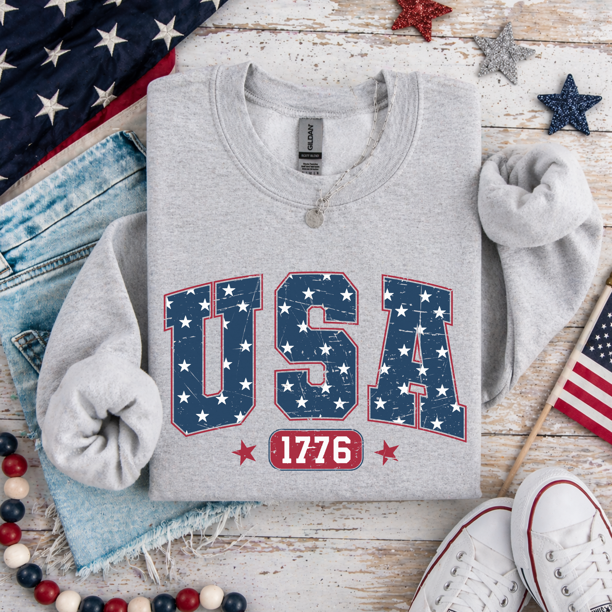 Gray sweatshirt with 'USA 1776' design on a wooden surface with American flags and jeans.