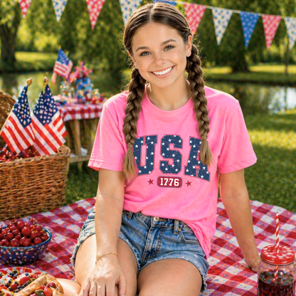 Young girl in a pink 'USA 1776' t-shirt sitting at a picnic table with American flags and food.