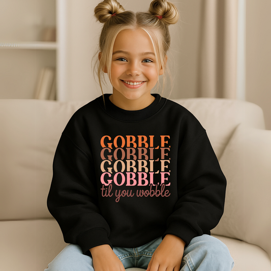 Youth girl wearing a black unisex Thanksgiving sweatshirt with colorful "Gobble Til You Wobble" text in orange, peach, and pink. Perfect fall outfit for girls, tweens, and kids. Cozy pullover ideal for turkey day, school parties, and family photos.
