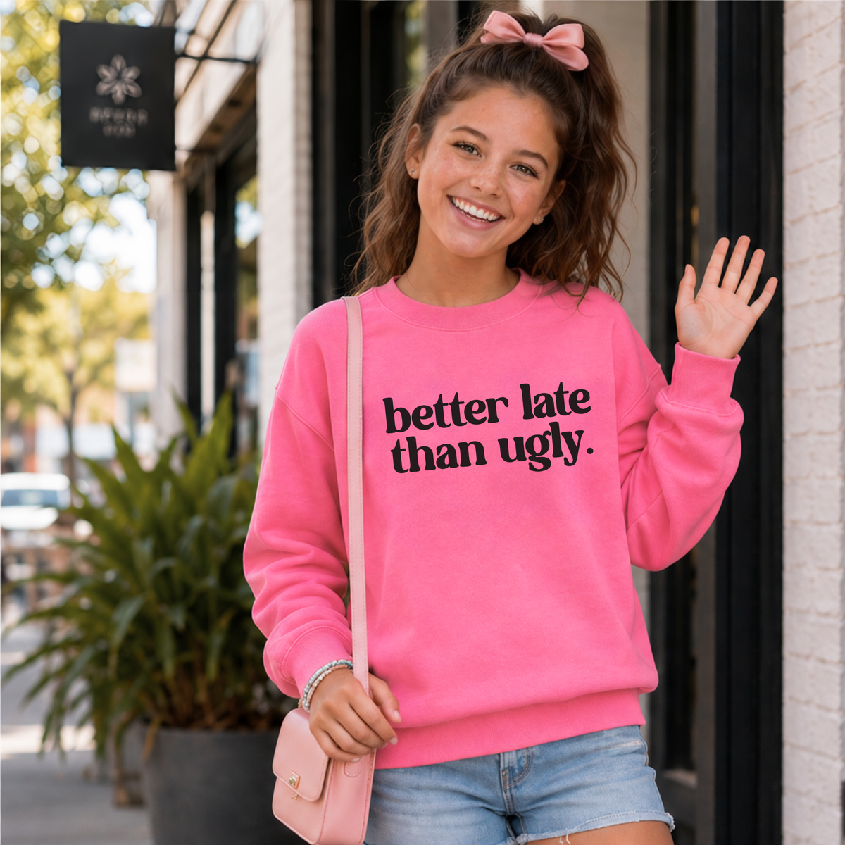 Young girl wearing a bright pink Better Late Than Ugly crewneck sweatshirt with black lettering outside a modern storefront.