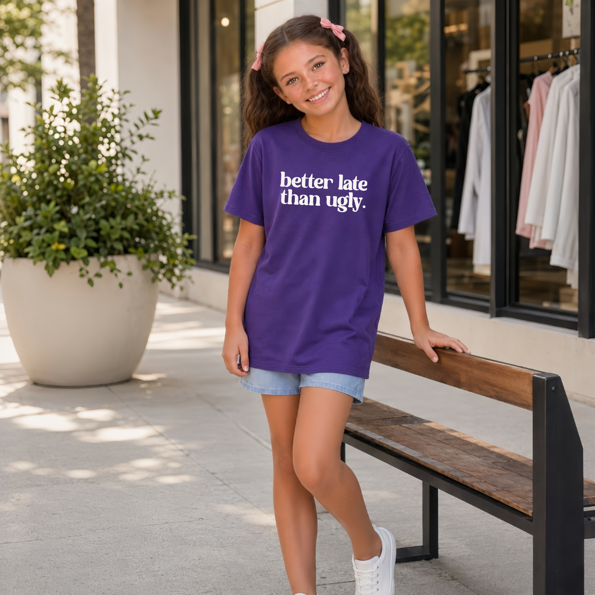Young girl wearing a purple short-sleeve Better Late Than Ugly T-shirt with white lettering outside a modern storefront.