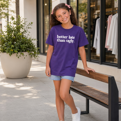 Young girl wearing a purple short-sleeve Better Late Than Ugly T-shirt with white lettering outside a modern storefront.