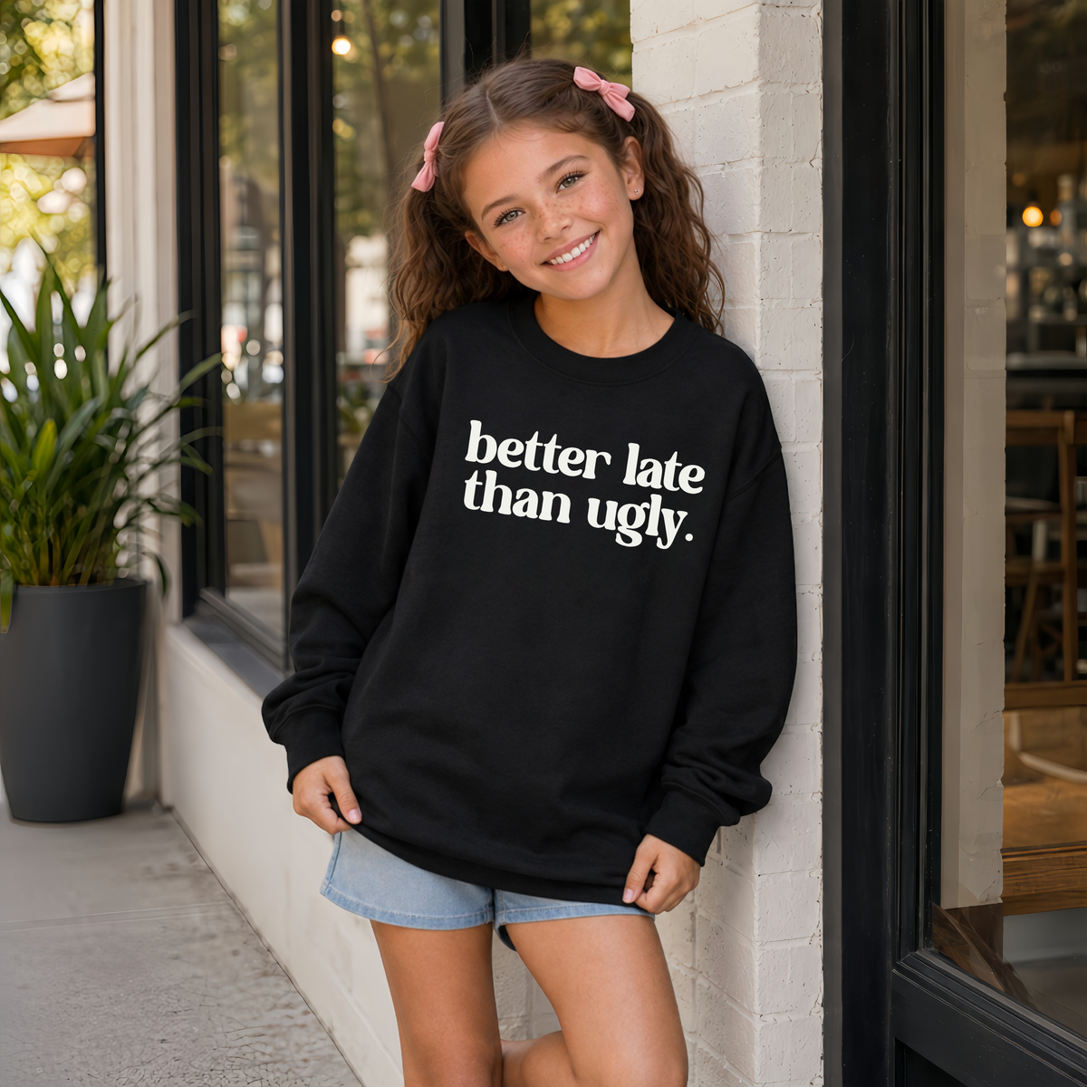 Young girl wearing a black Better Late Than Ugly crewneck sweatshirt with white lettering outside a modern storefront.