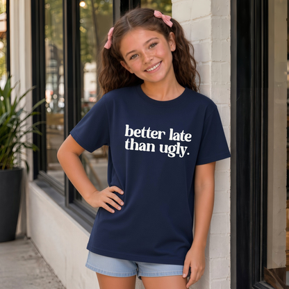 Young girl wearing a navy short-sleeve Better Late Than Ugly T-shirt with white lettering outside a modern storefront.