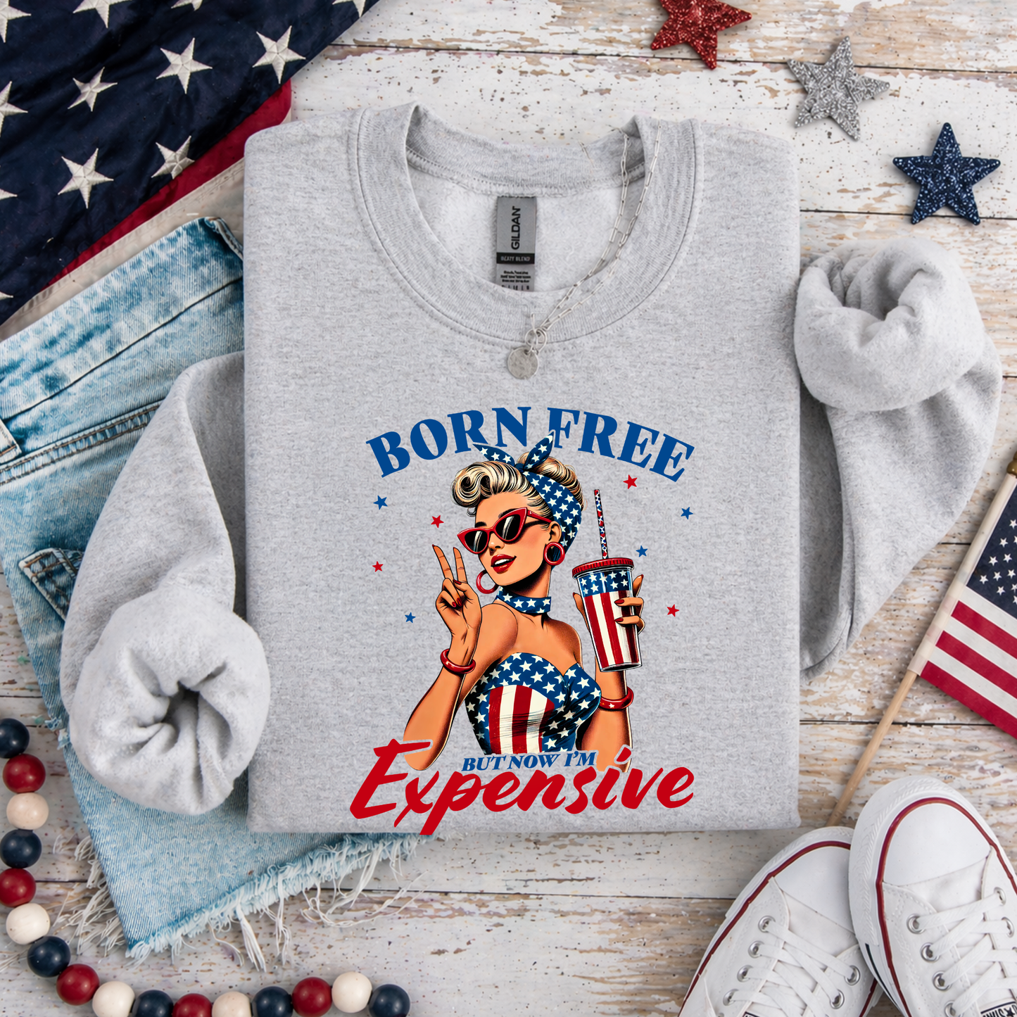 Gray sweatshirt with 'Born Free But Now I'm Expensive' graphic on a wooden surface with American flags and denim jeans.