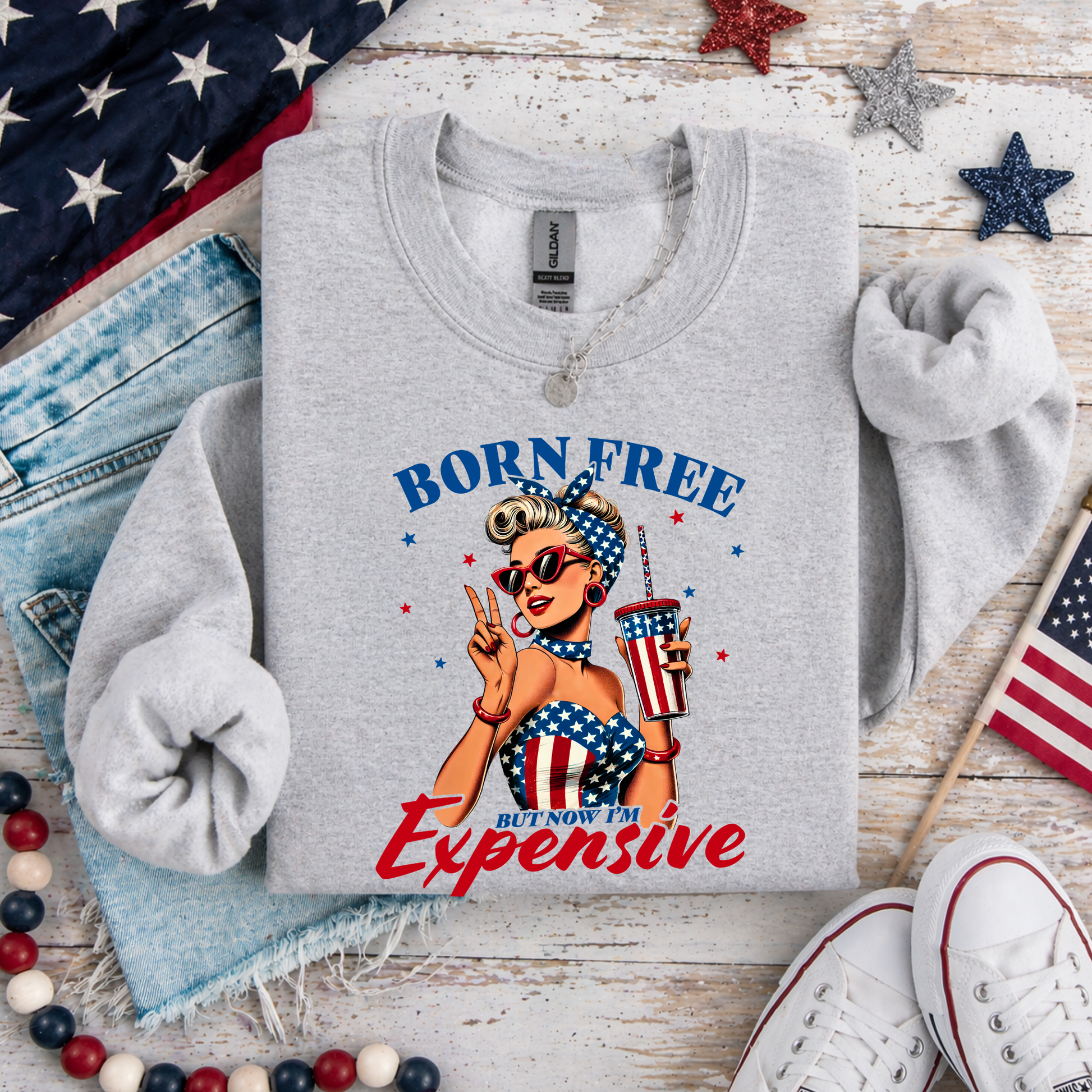 Gray sweatshirt with 'Born Free But Now I'm Expensive' graphic on a wooden surface with American flags and denim jeans.