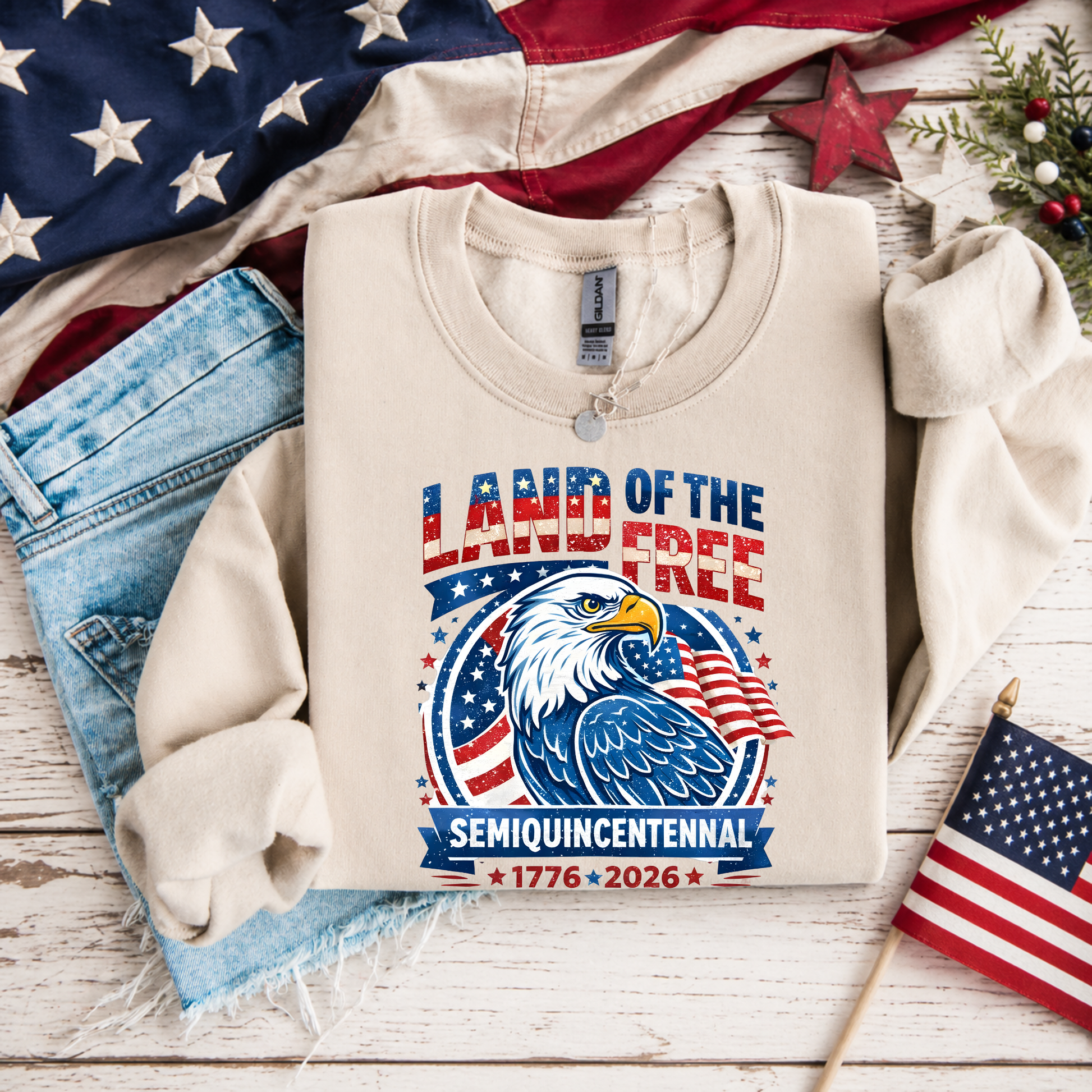 Beige sweatshirt with 'Land of the Free' design featuring an eagle and American flag, surrounded by American flags and denim jeans on a wooden surface.