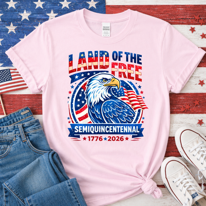 Pink t-shirt with eagle graphic and 'Land of the Free' text on a wooden surface with American flag background.