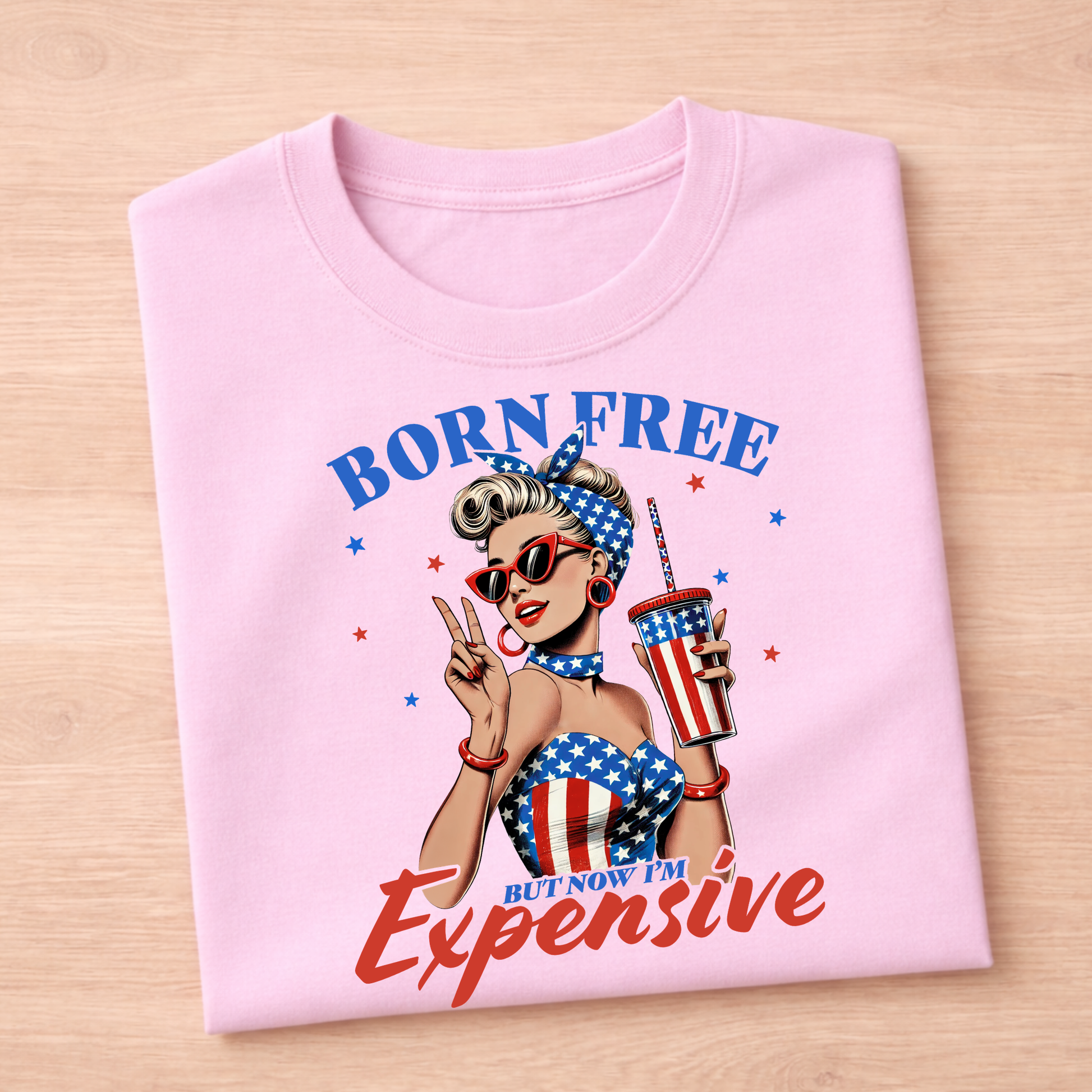Pink t-shirt with a graphic design of a woman holding a cup and sunglasses, with text 'Born Free But Now I'm Expensive' on a wooden surface.
