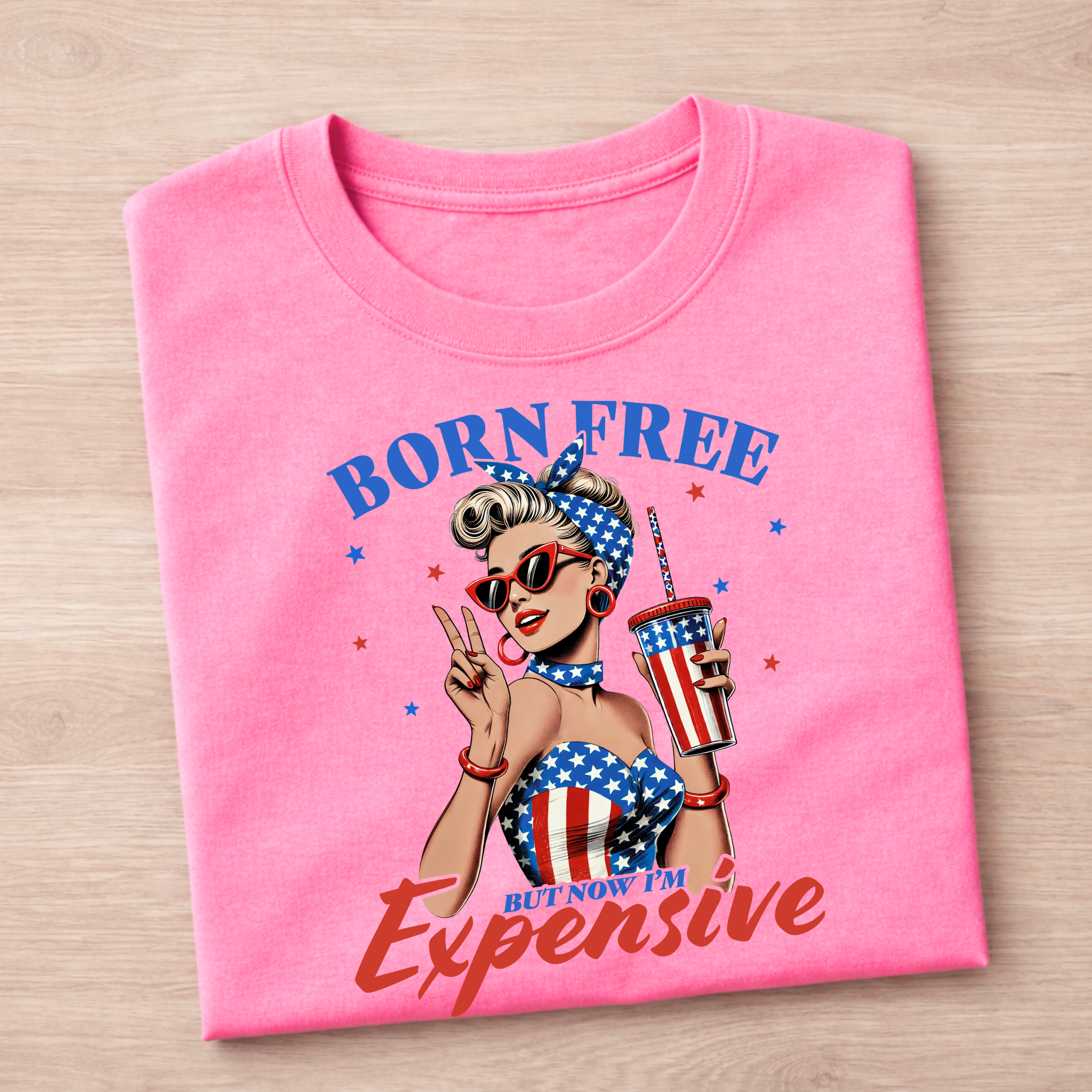Pink t-shirt with graphic design of a woman holding a cup and text 'Born Free But Now I'm Expensive' on a wooden surface.