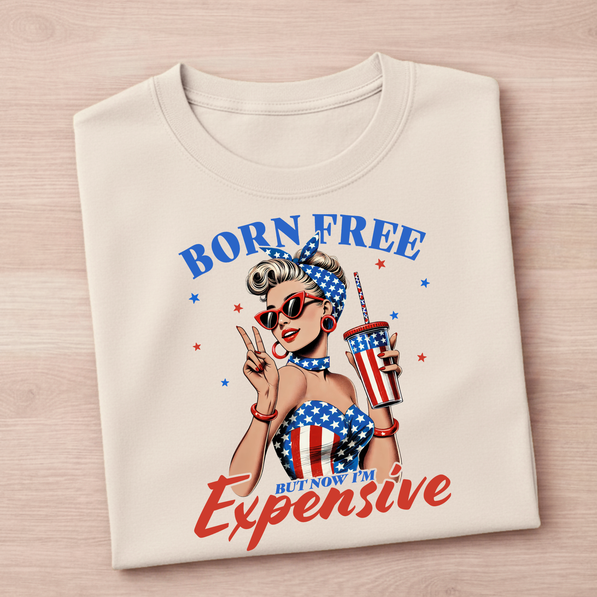 Folded t-shirt with a graphic design of a woman holding a drink, wearing an American flag outfit, and text 'Born Free But Now I'm Expensive' on a wooden surface.