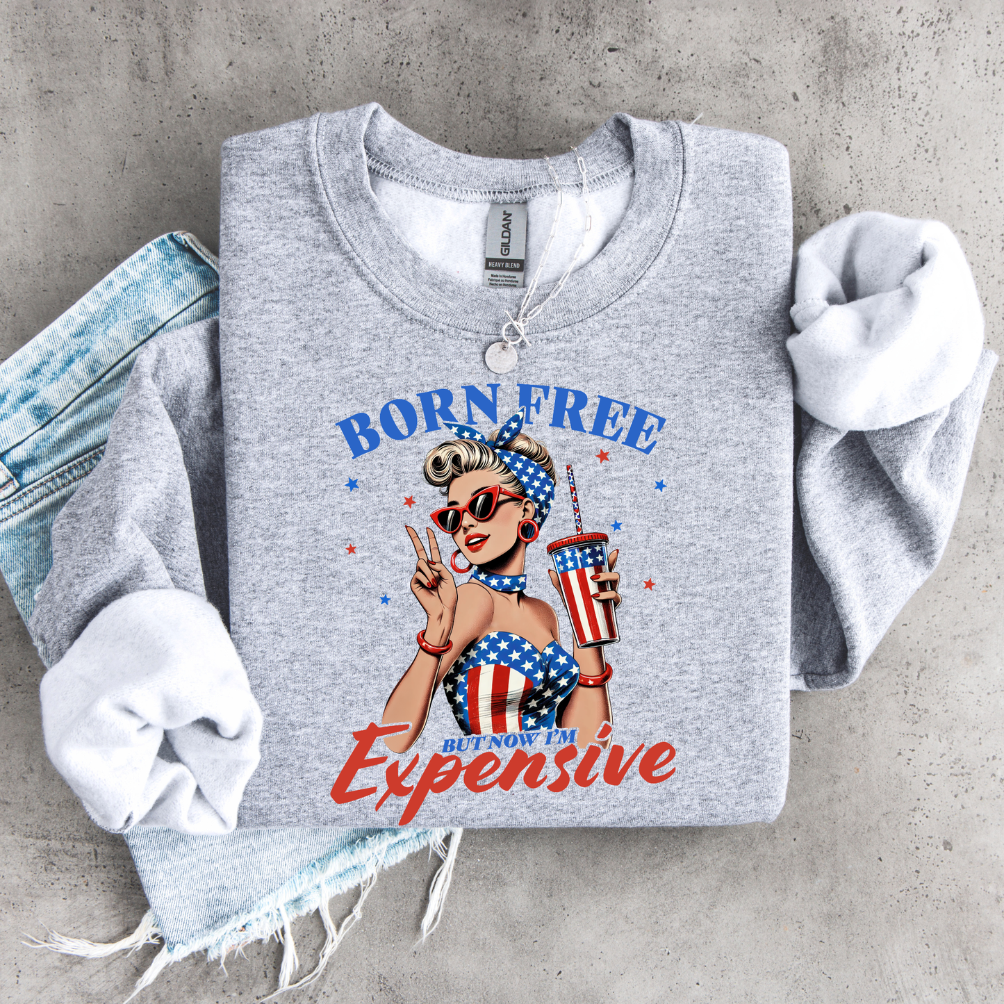 Gray sweatshirt with a graphic retro design of a woman in a patriotic outfit and text 'Born Free But Now I'm Expensive' on a concrete surface.