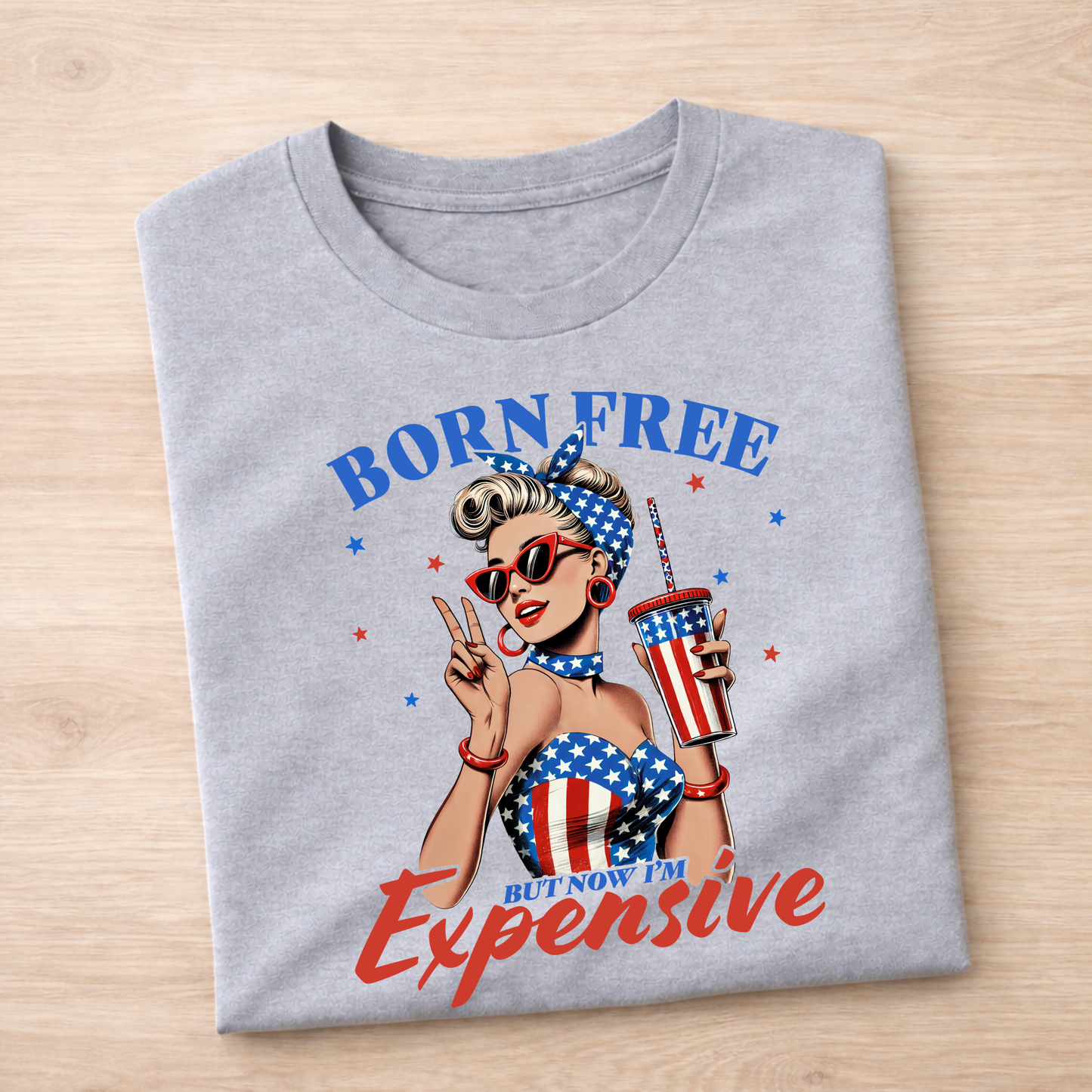 Folded gray t-shirt with a graphic design of a woman holding a cup and sunglasses, with text 'Born Free But Now I'm Expensive' on a wooden surface.