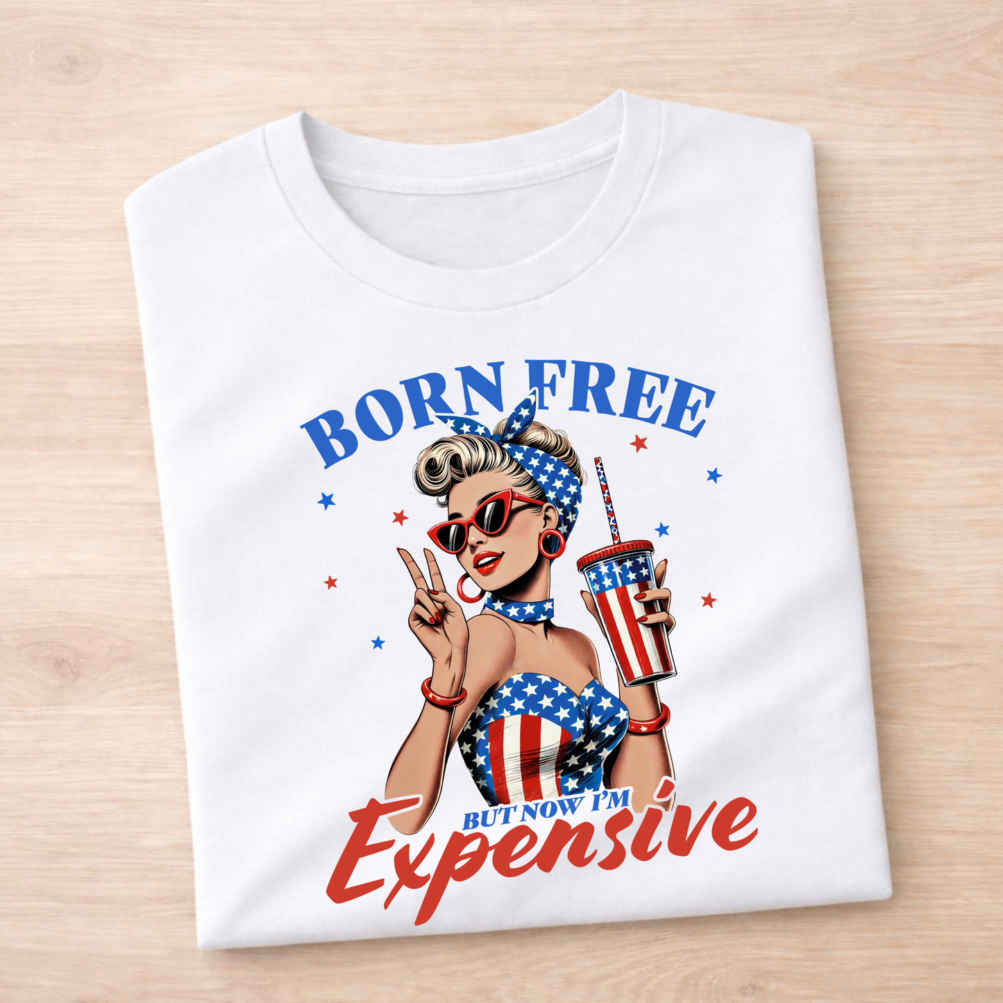 White t-shirt with a graphic design of a woman holding a drink, sunglasses, and an American flag-themed outfit on a wooden surface.