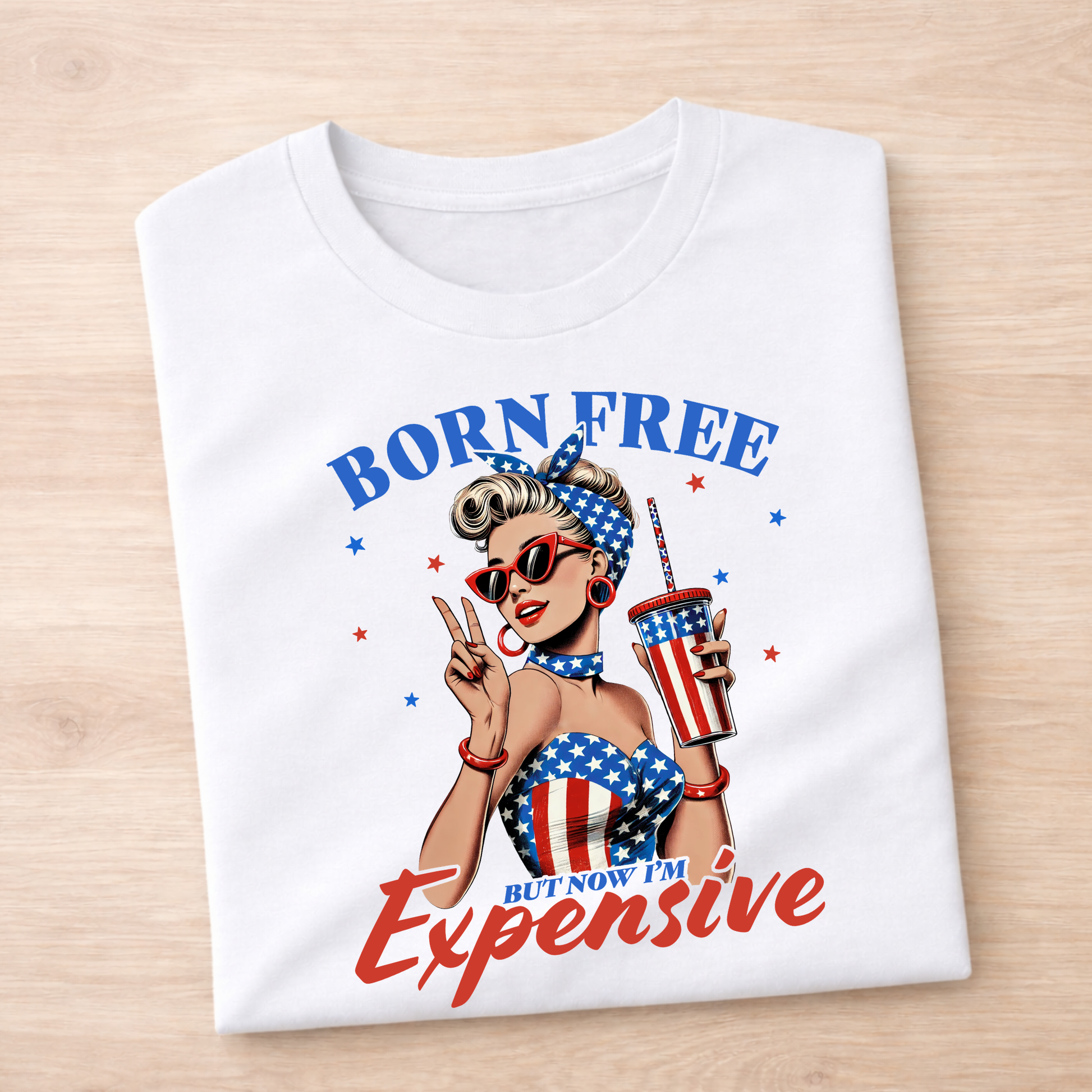 White t-shirt with a graphic design of a woman holding a drink, sunglasses, and an American flag-themed outfit on a wooden surface.