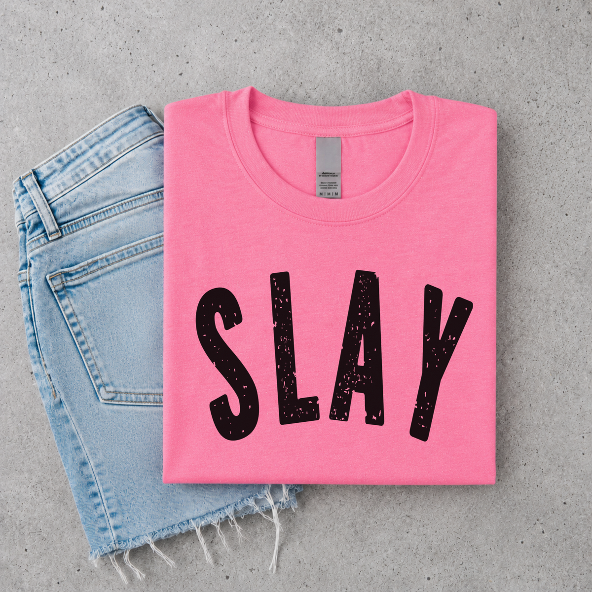 Bright pink SLAY short-sleeve T-shirt flat lay with bold black distressed lettering, styled with denim.
