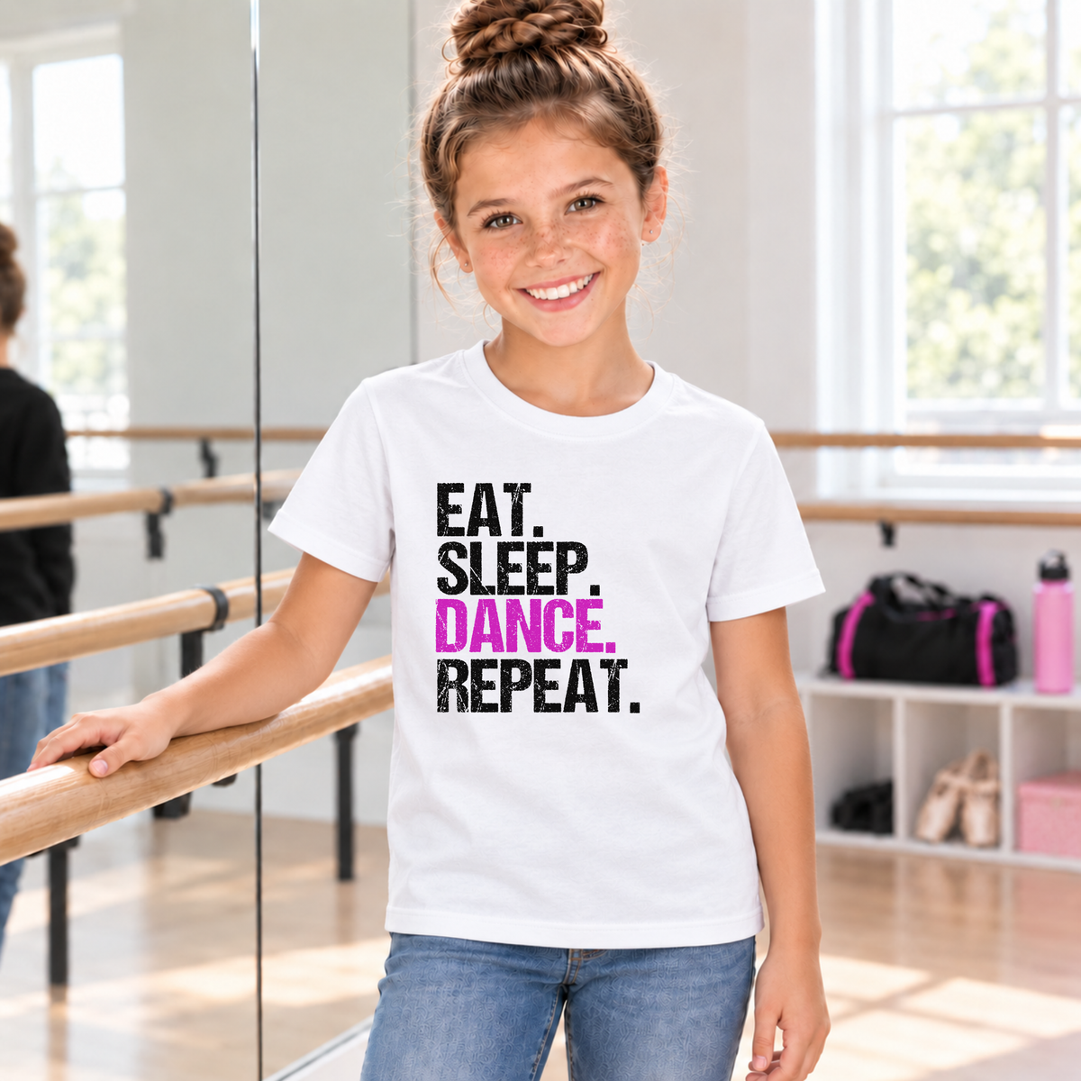 Girl wearing a white youth Eat Sleep Dance Repeat T-shirt beside a ballet barre in a sunlit studio.