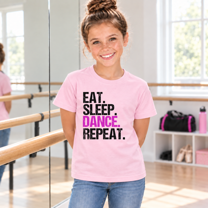 Girl in a light pink youth Eat Sleep Dance Repeat T-shirt standing near a ballet barre and mirror.