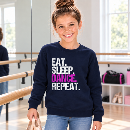 Girl wearing a navy youth Eat Sleep Dance Repeat sweatshirt while holding ballet shoes in a dance studio.