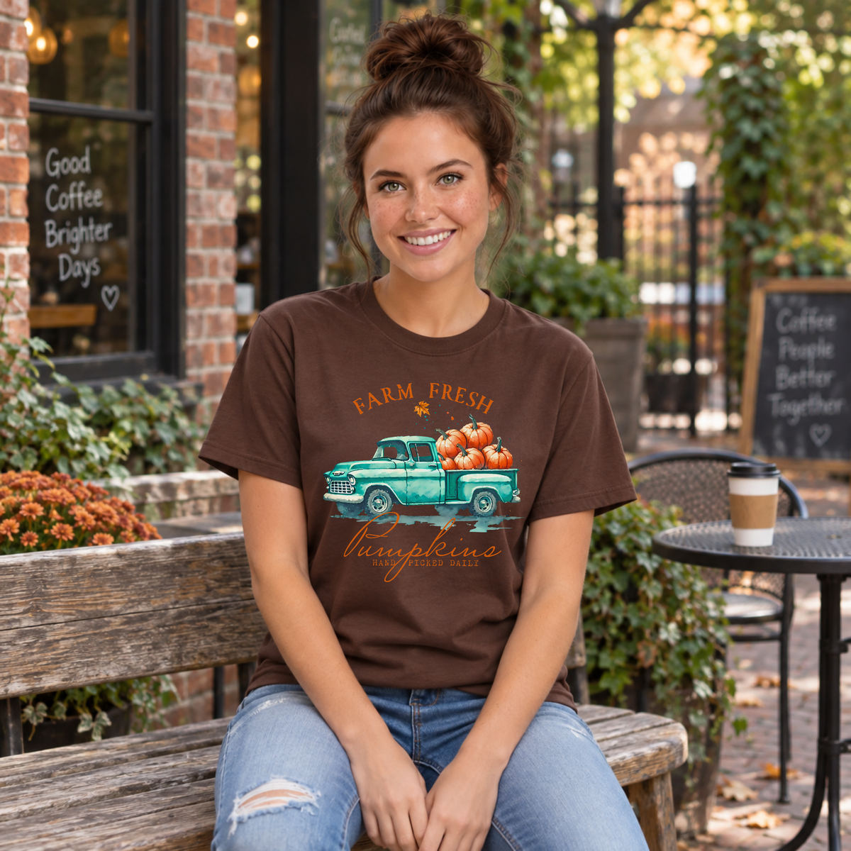 Woman in a brown T-shirt with a teal truck full of pumpkins and Farm Fresh Pumpkins design seated outside a fall cafe.