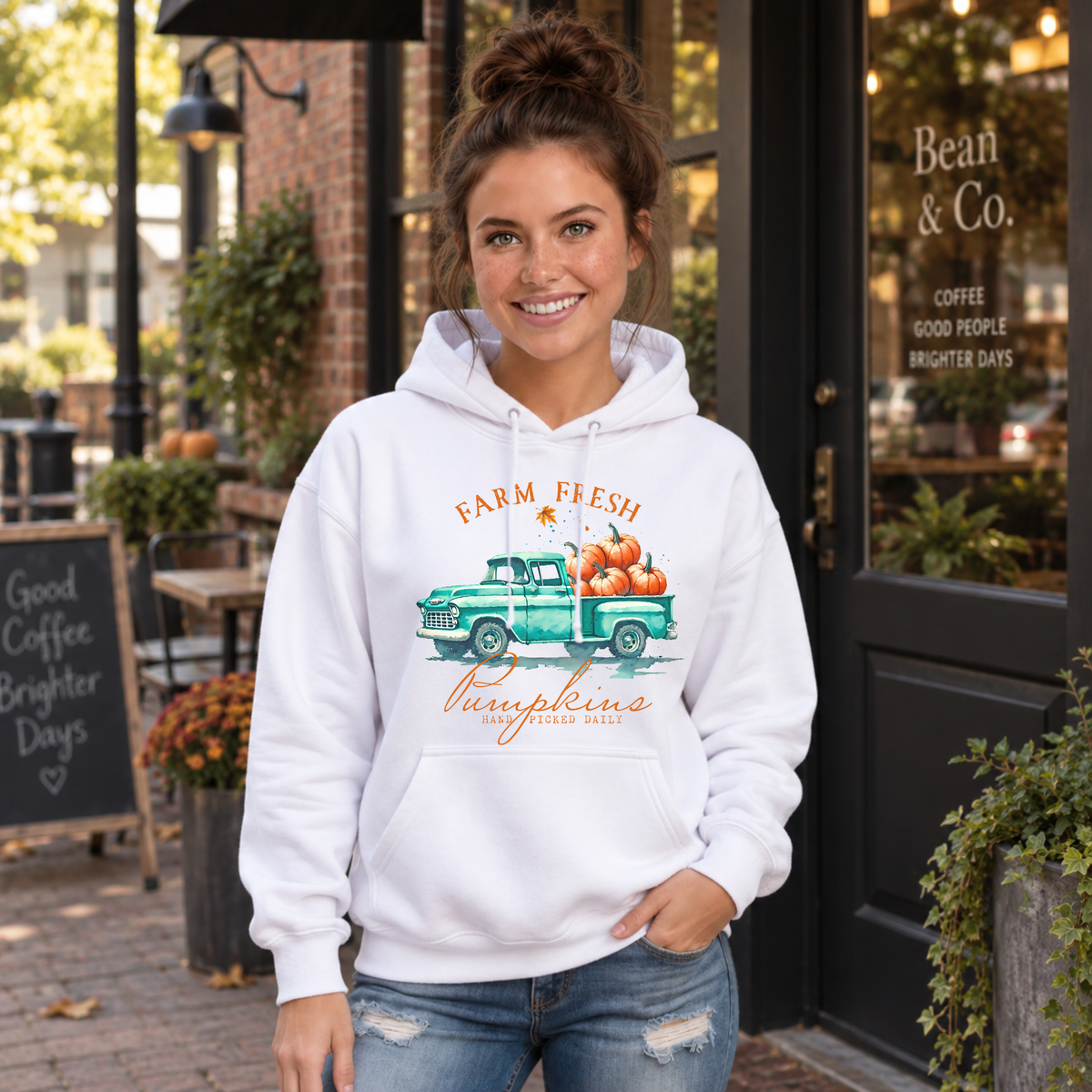 Woman wearing a white hoodie with a vintage teal pumpkin truck and Farm Fresh Pumpkins design outside a cafe.